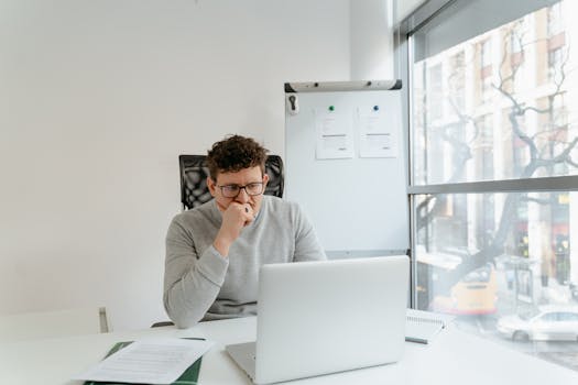 A thoughtful man in a gray sweater concentrates on his laptop at a bright office desk.
